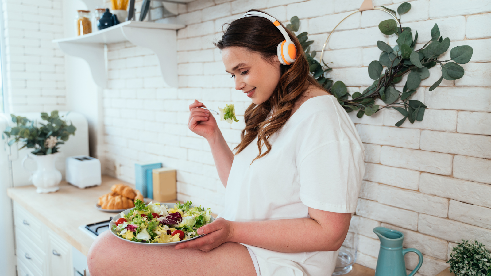 A woman sitting on a counter with headphones on eating healthy meal.