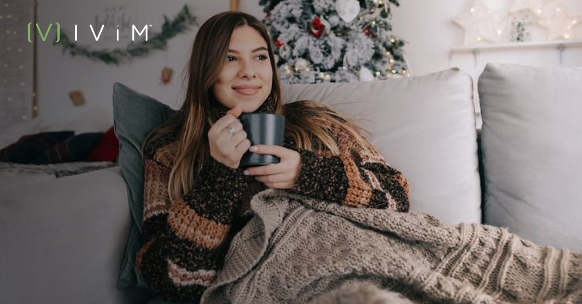 Woman relaxing on couch with a blanket and mug, surrounded by holiday decorations, promoting stress-free holiday wellness.